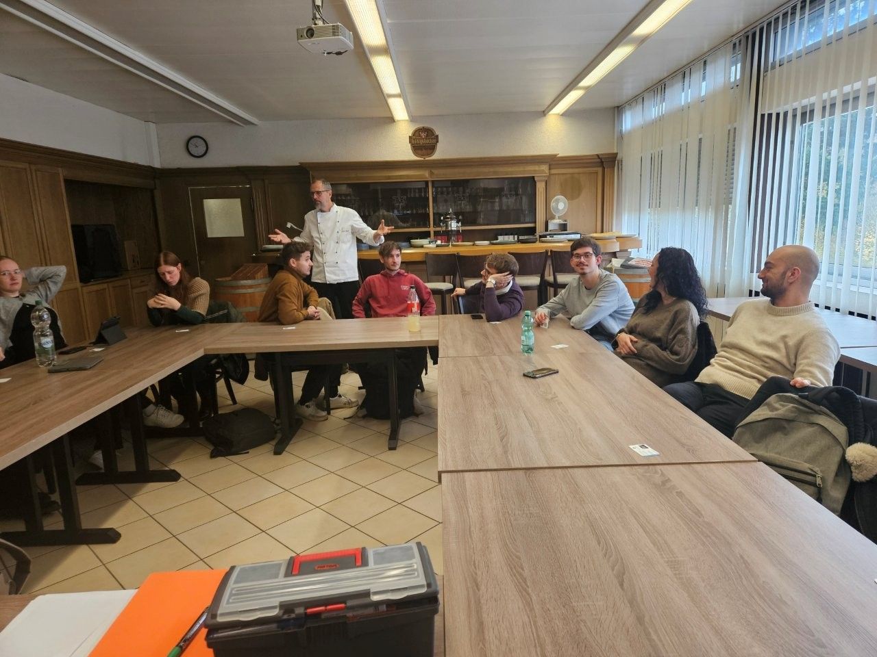 Menschen sitzen an einem Tisch in einem Raum und hören einem Mann zu, der spricht und dabei mit den Händen gestikuliert.