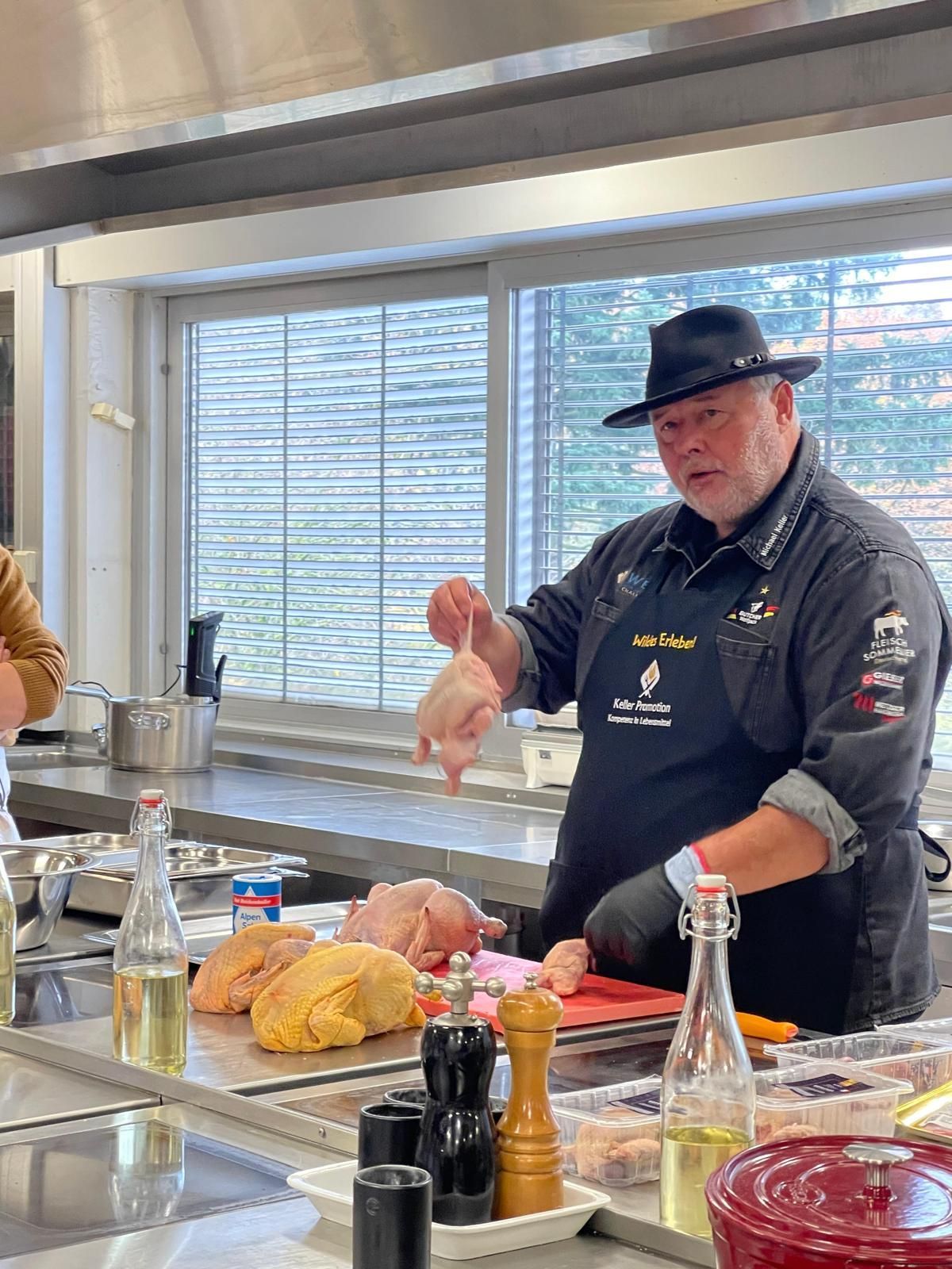 Ein Koch mit schwarzer Kochmütze demonstriert das Zerlegen von Hühnern in einer Küche.
