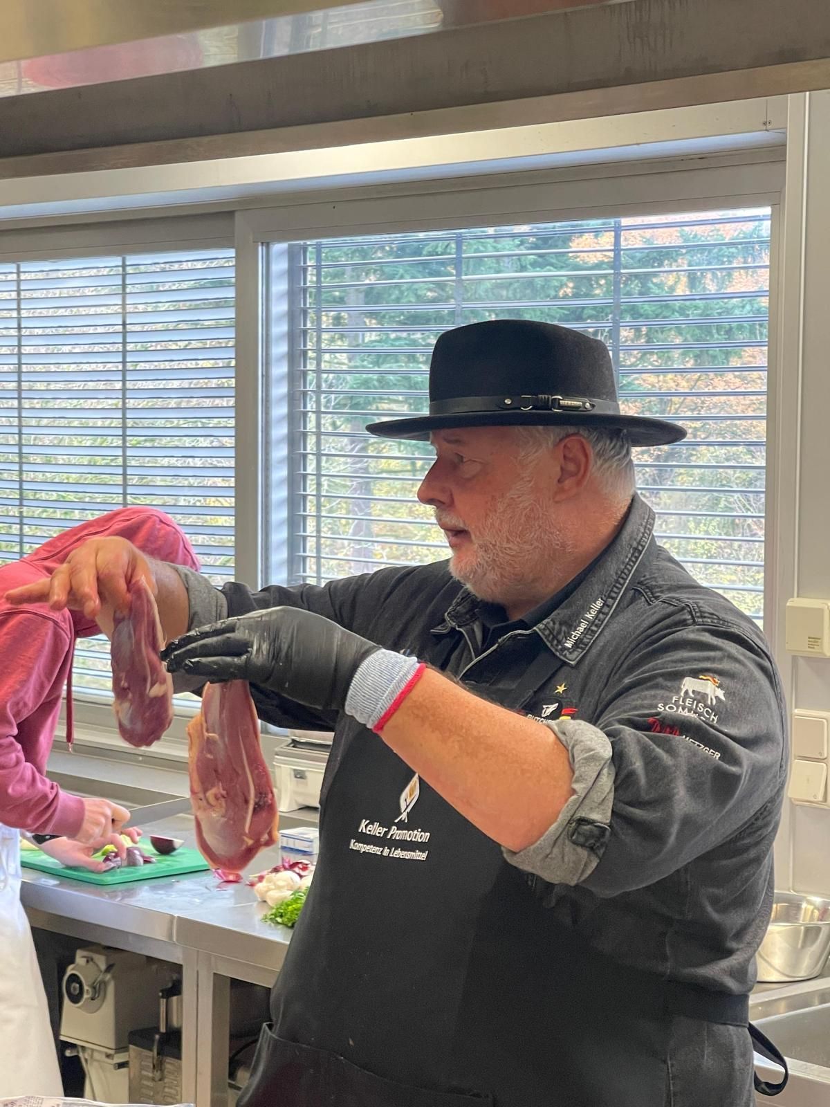 Ein Mann mit schwarzem Hut und Schürze hält rohes Fleisch in einer Küche.