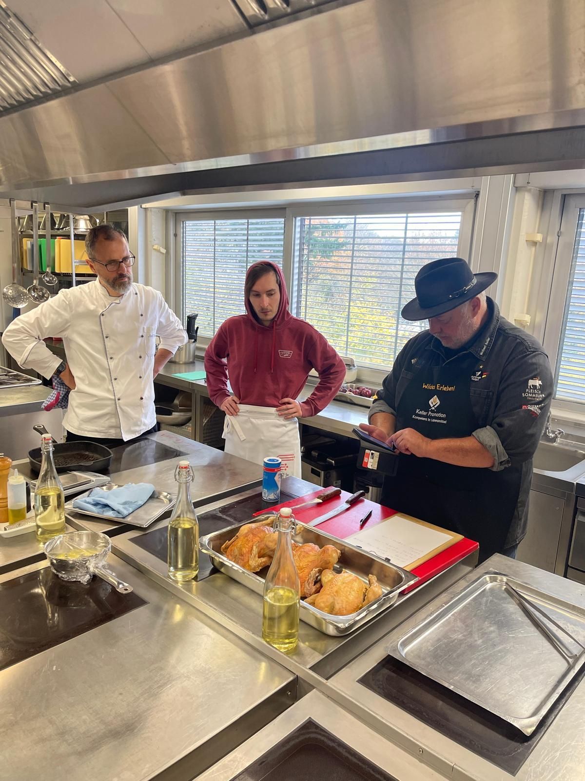 Köche bereiten in einer Küche Speisen zu; einer schneidet, während die anderen zusehen. Sonnenlicht strömt durch ein Fenster herein.