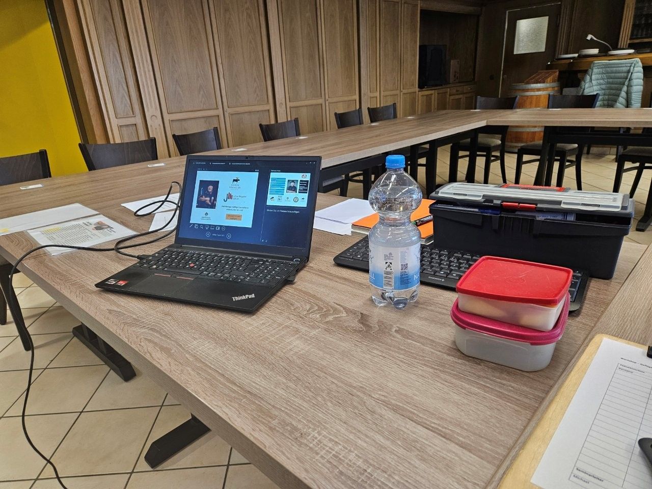 Laptop auf einem langen Holztisch, daneben Wasserflasche, Tastatur und Behälter. Innenaufnahme.