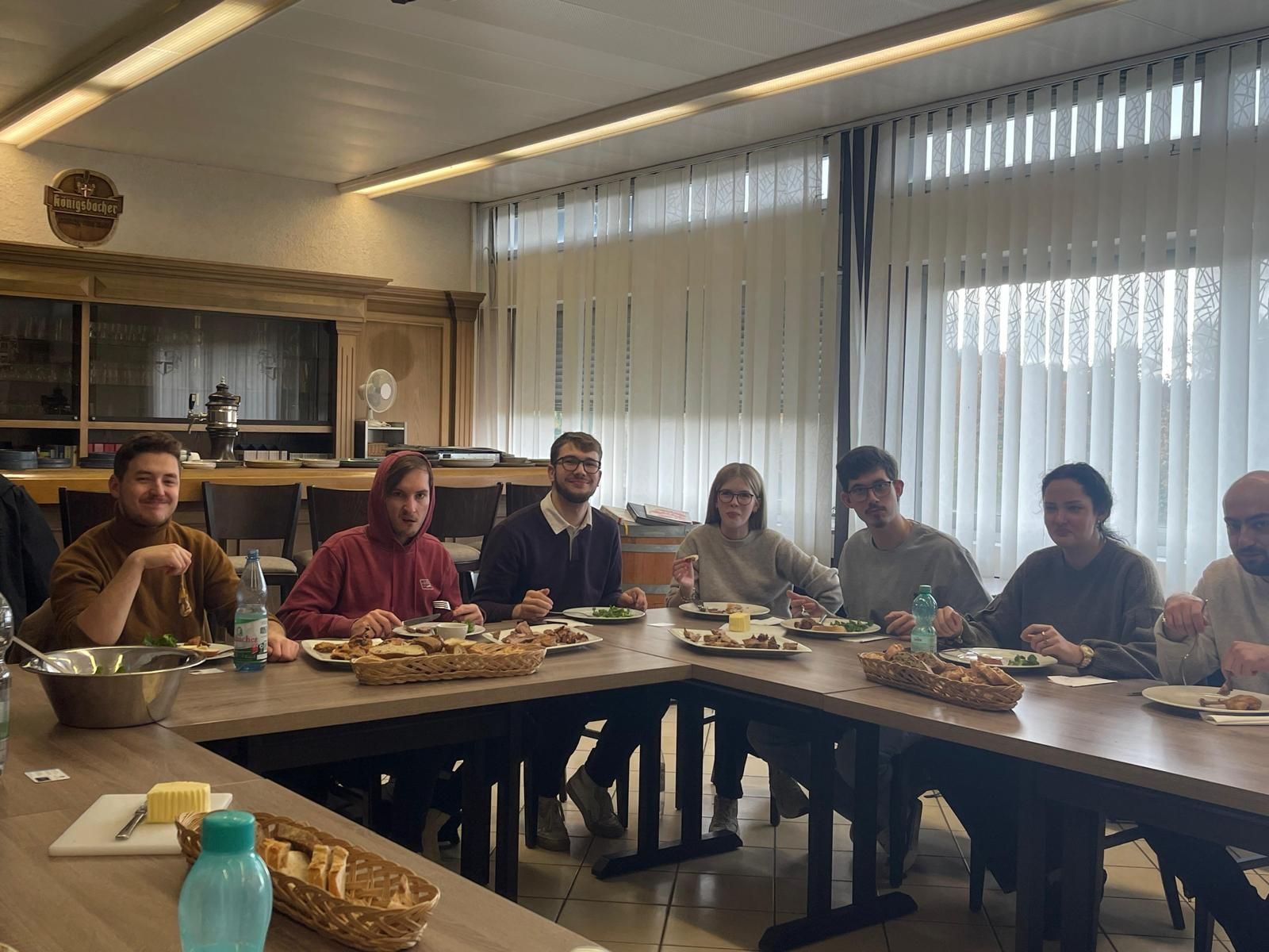 Menschen sitzen um einen Tisch und essen. Sie befinden sich in einem Innenraum mit einem großen Fenster und Holzvertäfelung.