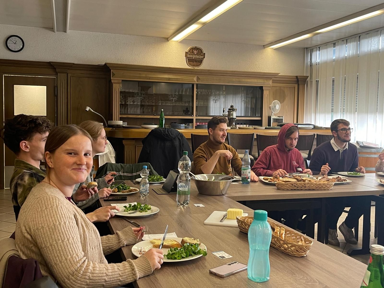 Menschen sitzen um einen Tisch und essen. Eine Person lächelt in die Kamera. Ein Zimmer mit Holzvertäfelung, einer Uhr und einem Fenster.