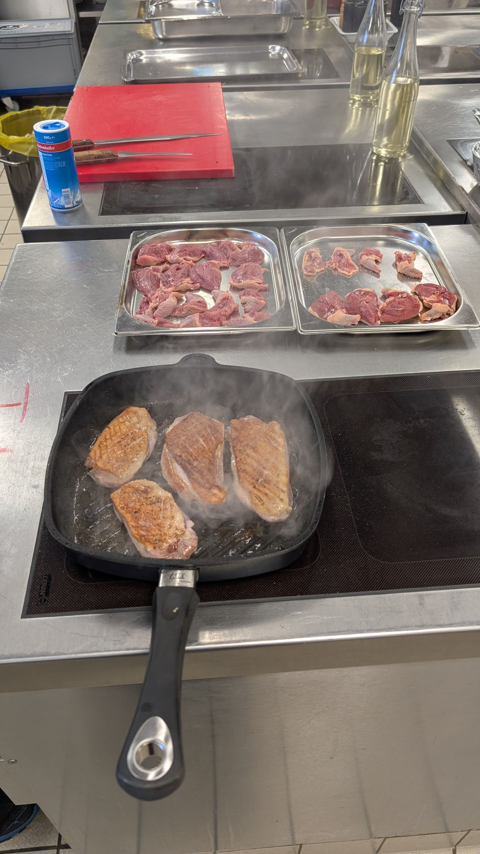 In einer professionellen Küche brät Fleisch in einer quadratischen Pfanne auf dem Herd, daneben stehen Behälter mit rohem Fleisch.
