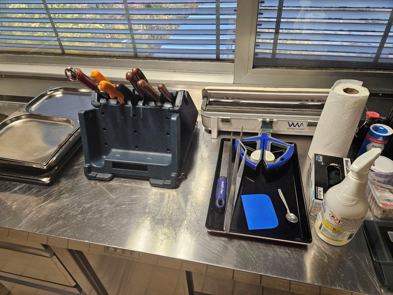 Arbeitsplatte aus Edelstahl mit Küchenutensilien: Messer im Halter, Tabletts, ein Versiegelungsgerät und andere Utensilien, in der Nähe eines Fensters.