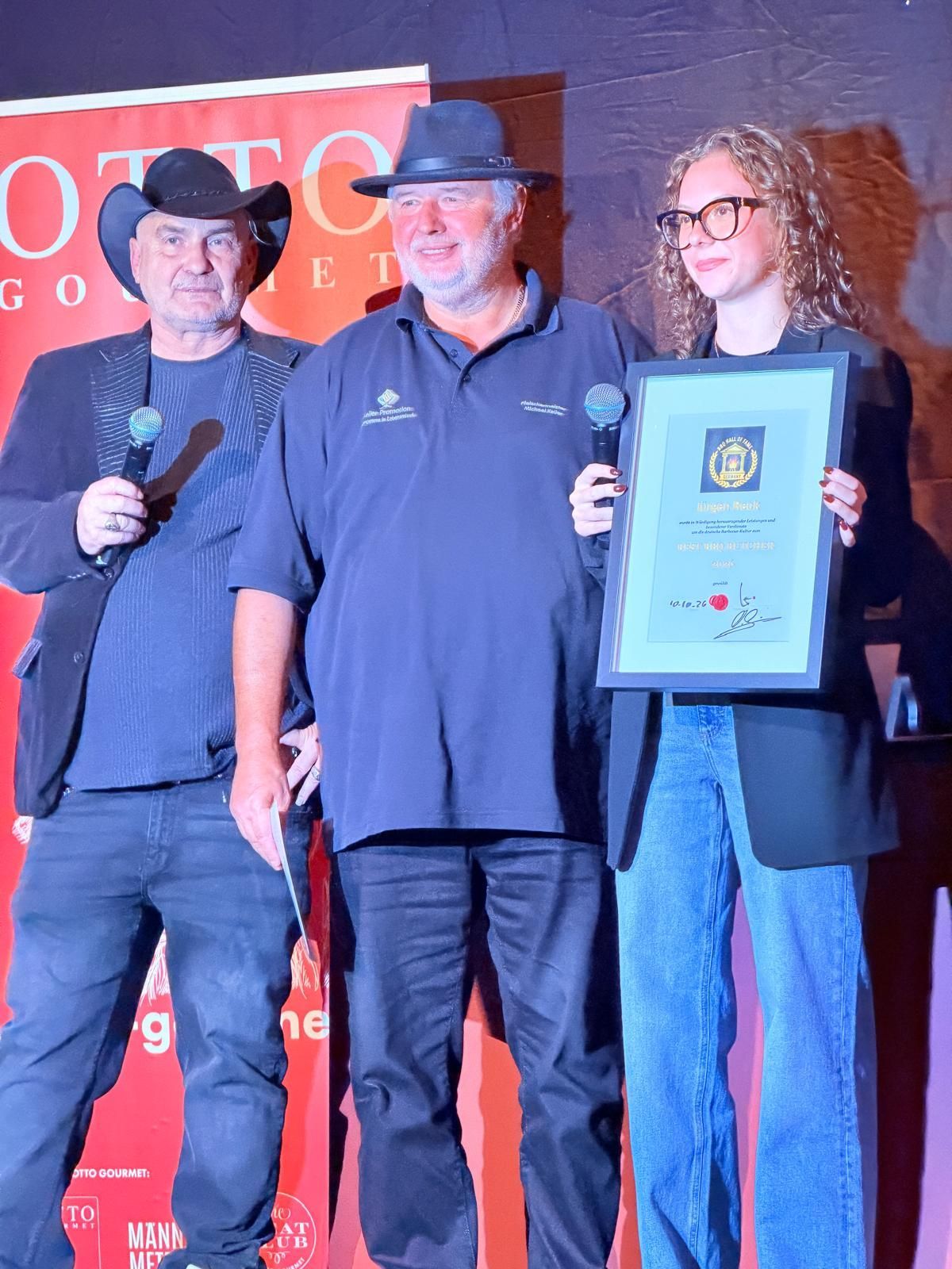 Three people on stage holding microphones and a framed award, with a red event banner behind them.