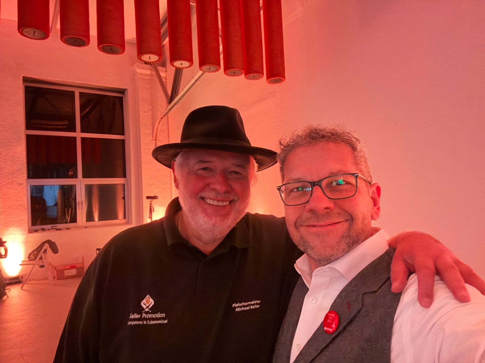 Two smiling men pose together in a warmly lit room, one in a black hat and one in a gray suit.