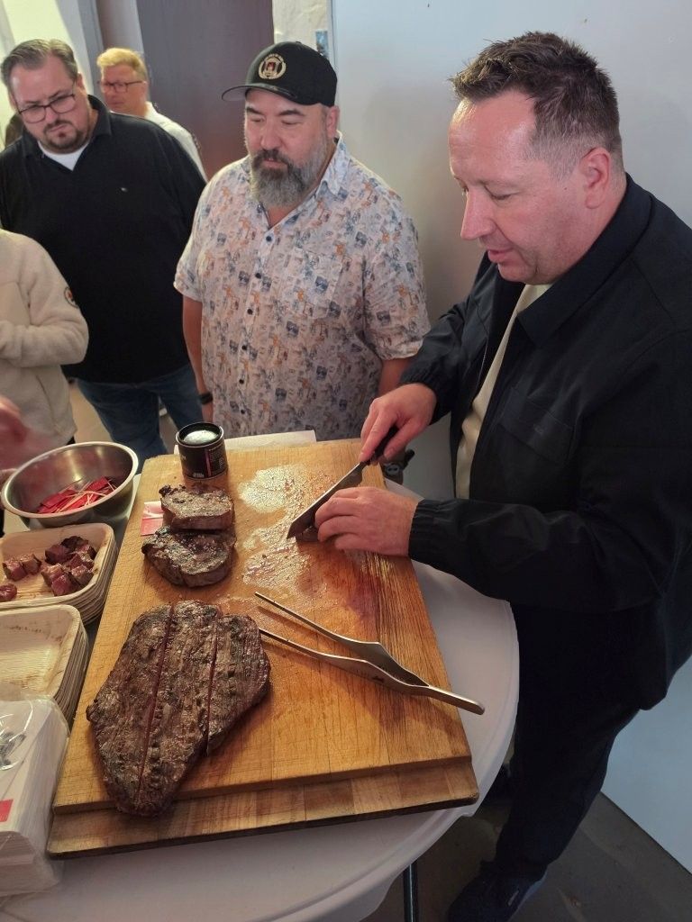 Three men at a table, one slicing a large roast on a wooden cutting board beside serving dishes.