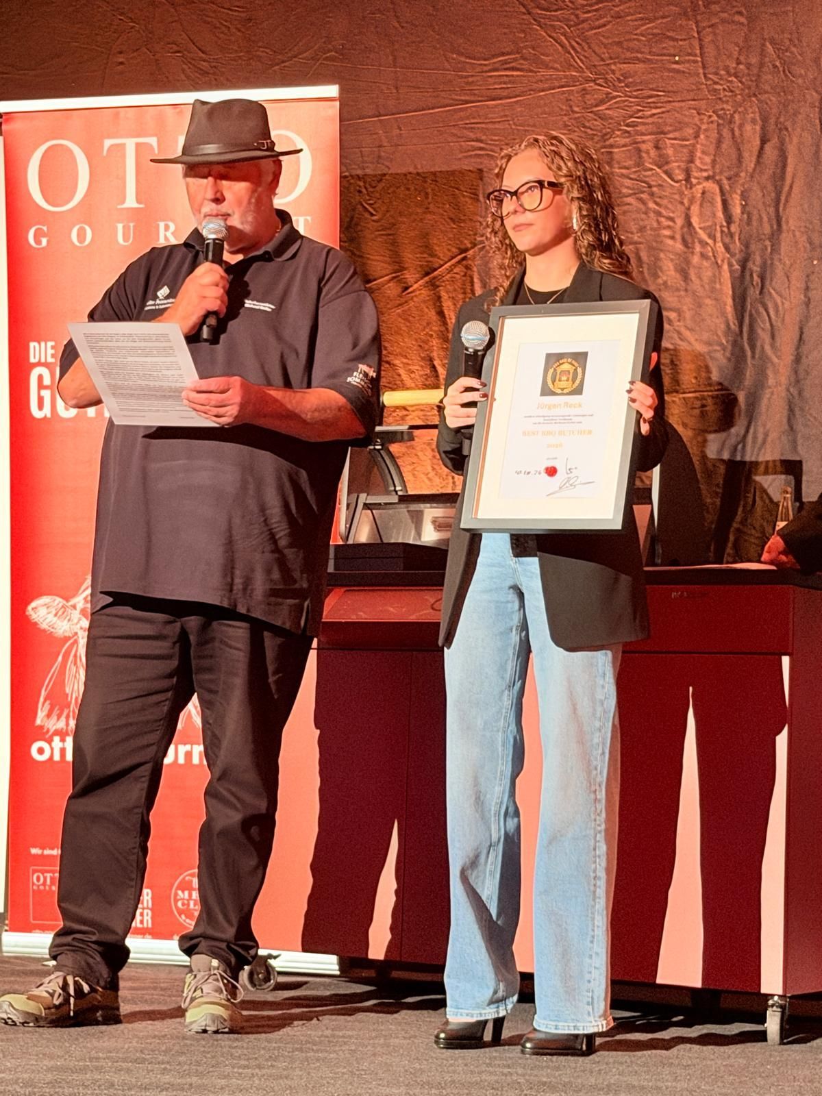 Two people onstage; one reads from a paper while the other holds a framed award certificate.