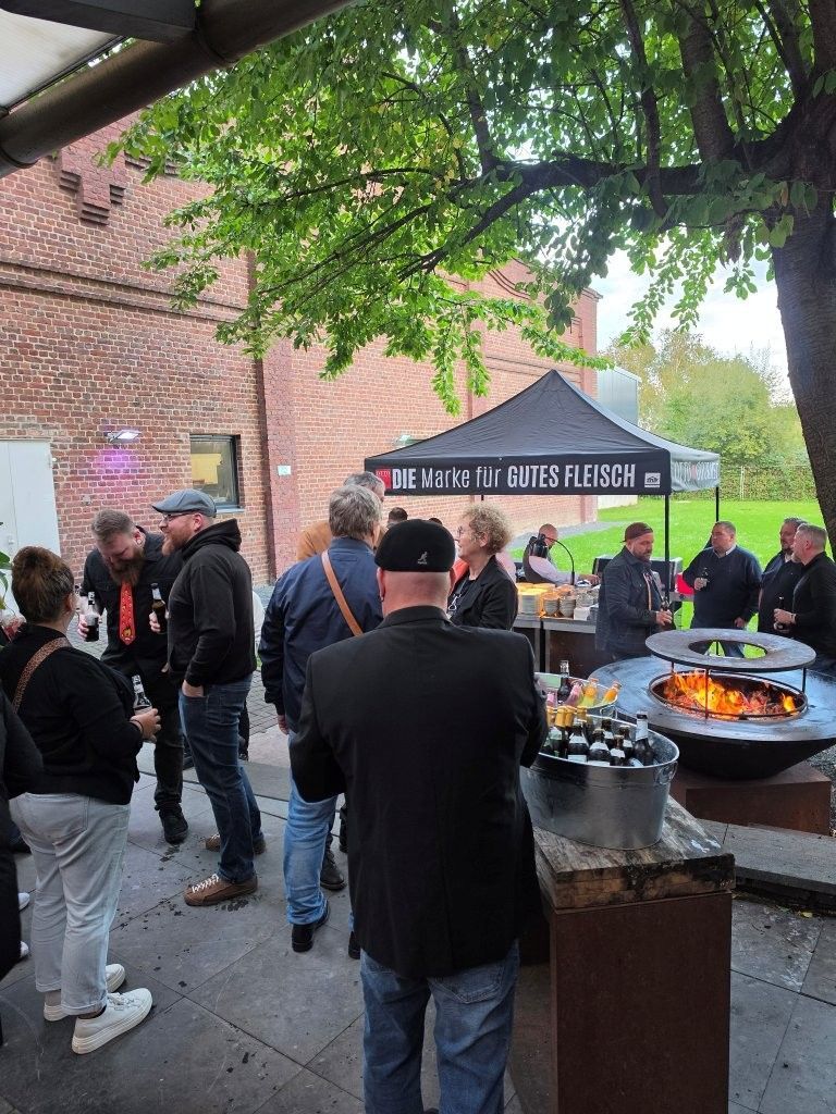 People gathered under a canopy at an outdoor grill, with flames visible and a brick wall in the background.
