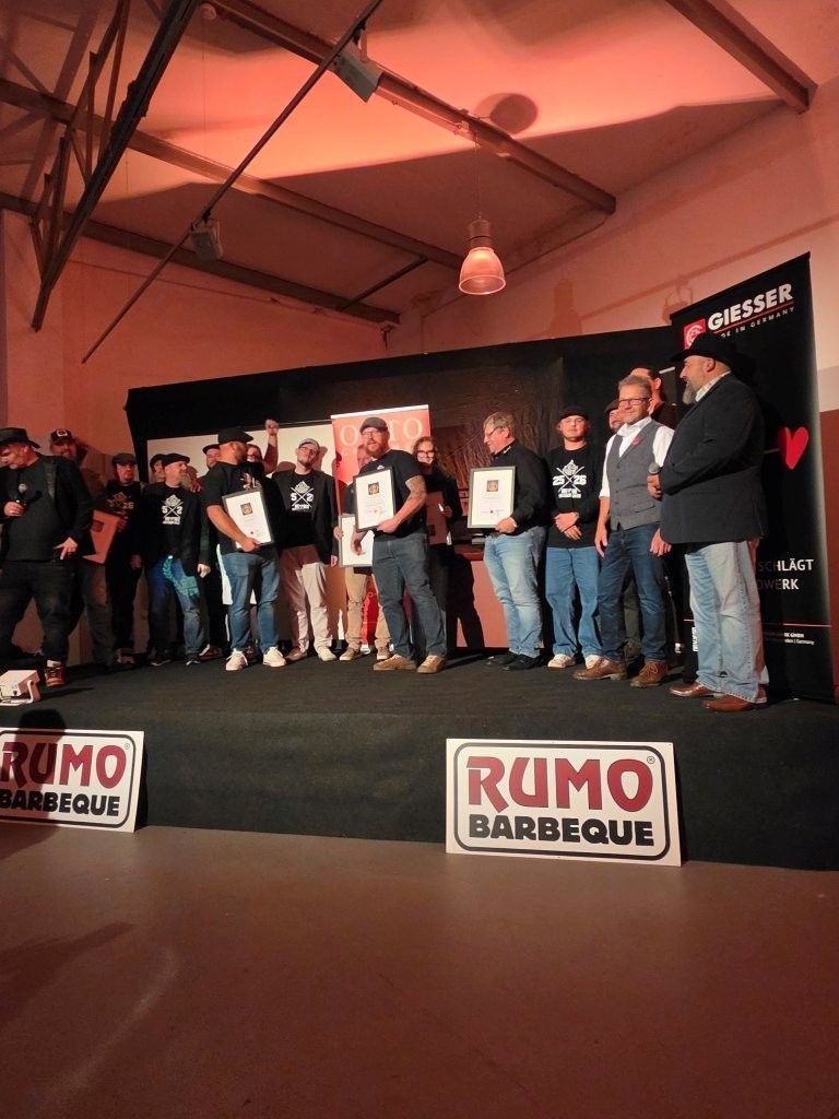 Group of people standing on a stage at a barbecue event with RUMO BBQ signs and sponsor banners