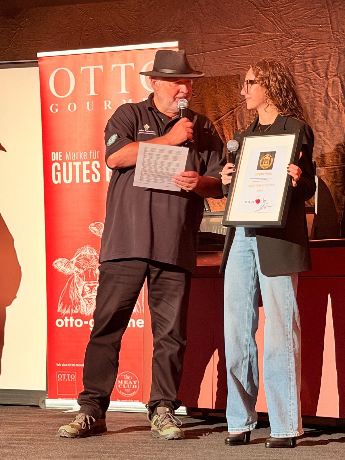 Two people onstage holding a certificate and plaque beside an OTTO banner.