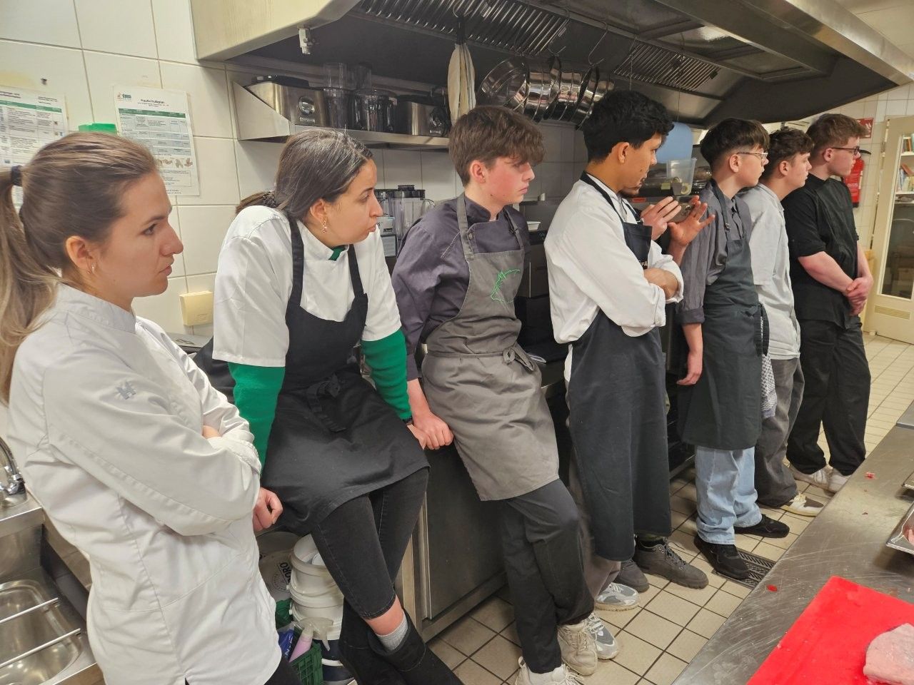 Köche in Schürzen lehnen an einer Theke in einer Großküche und beobachten eine Vorführung.