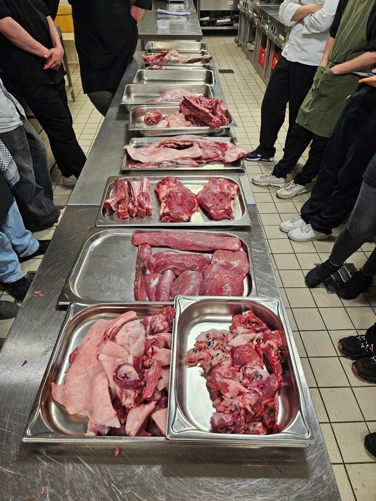 Rohes Fleisch wird in Tabletts auf einer Edelstahlfläche präsentiert, umgeben von Menschen in einer Küche.