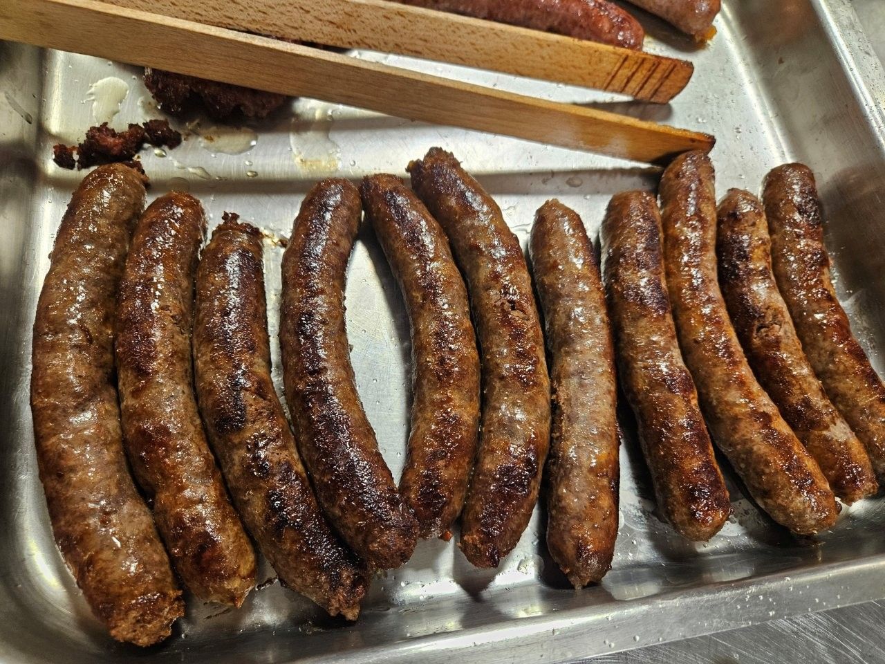 Gegrillte Würstchen in einer Metallschale, darauf eine Holzzange.