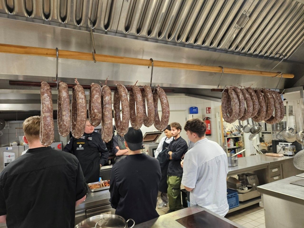 Würstchen hängen in einer Küche; eine Gruppe von Menschen beobachtet sie, einige in Kochkleidung.