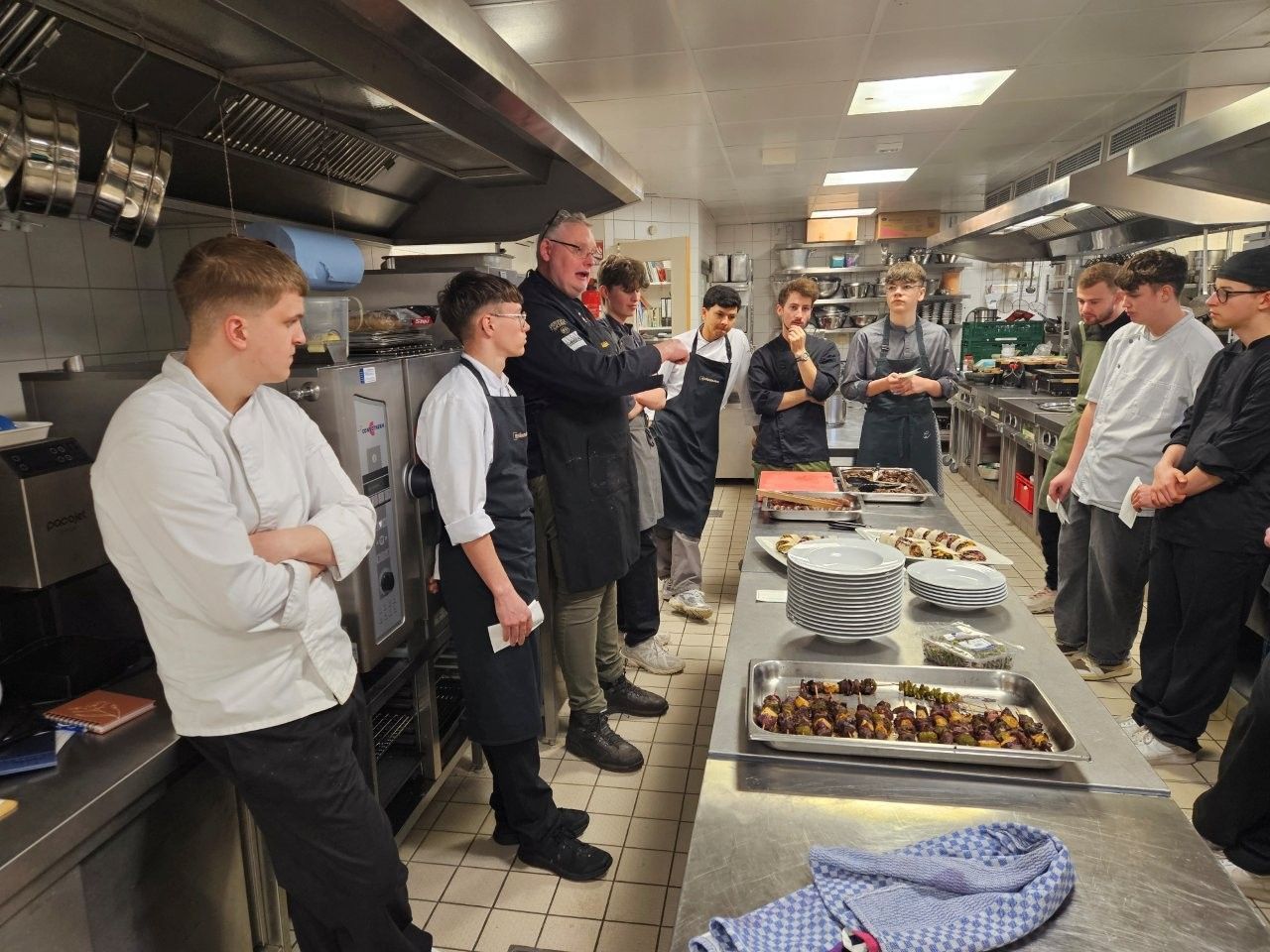 Ein Küchenchef unterrichtet Kochschüler in einer Großküche. Die Schüler beobachten die Zubereitung von Speisen.
