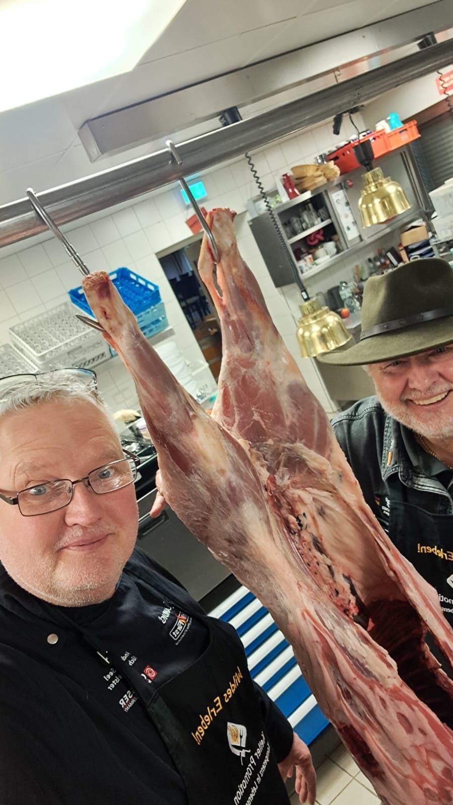 Zwei Männer posieren mit einem hängenden Tierkadaver in einer Küche. Einer trägt Hut und Schürze; beide lächeln.