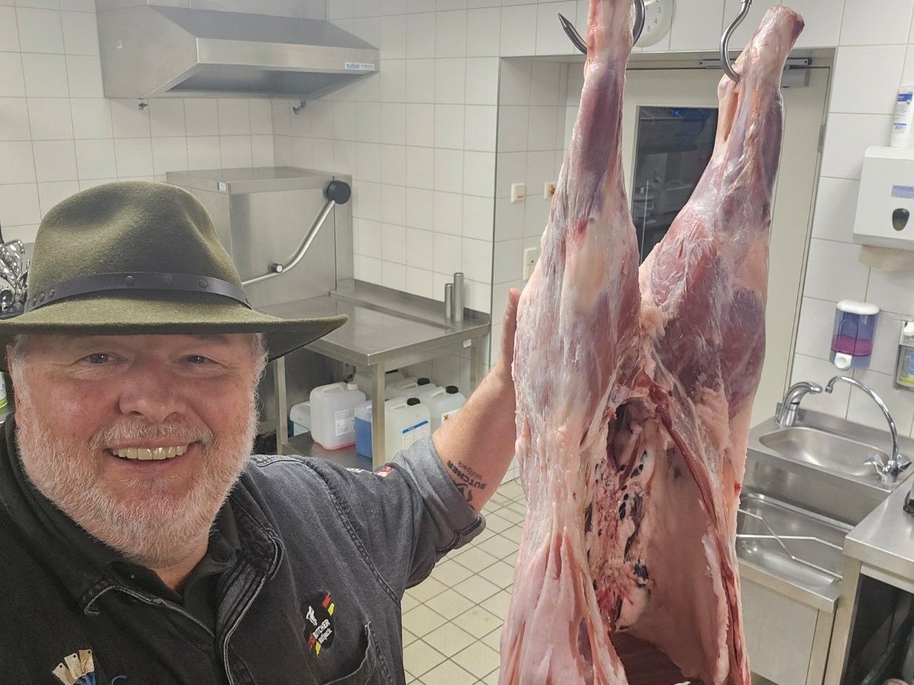 Ein lächelnder Mann mit Hut hält einen in einer Küche aufgehängten, geschlachteten Tierkadaver in der Hand.