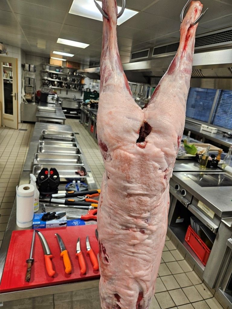 Ein ganzer Lammkörper hängt in einer Großküche neben Metzgermessern auf einem roten Schneidebrett.