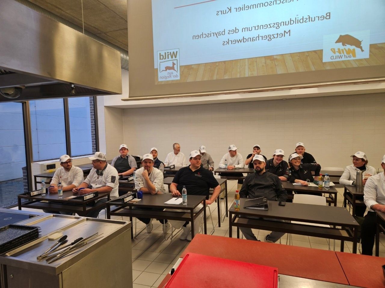 Köche mit weißen Kochmützen bei einem Kochkurs, die in einem hellen Raum mit Präsentationsbildschirm Notizen machen.