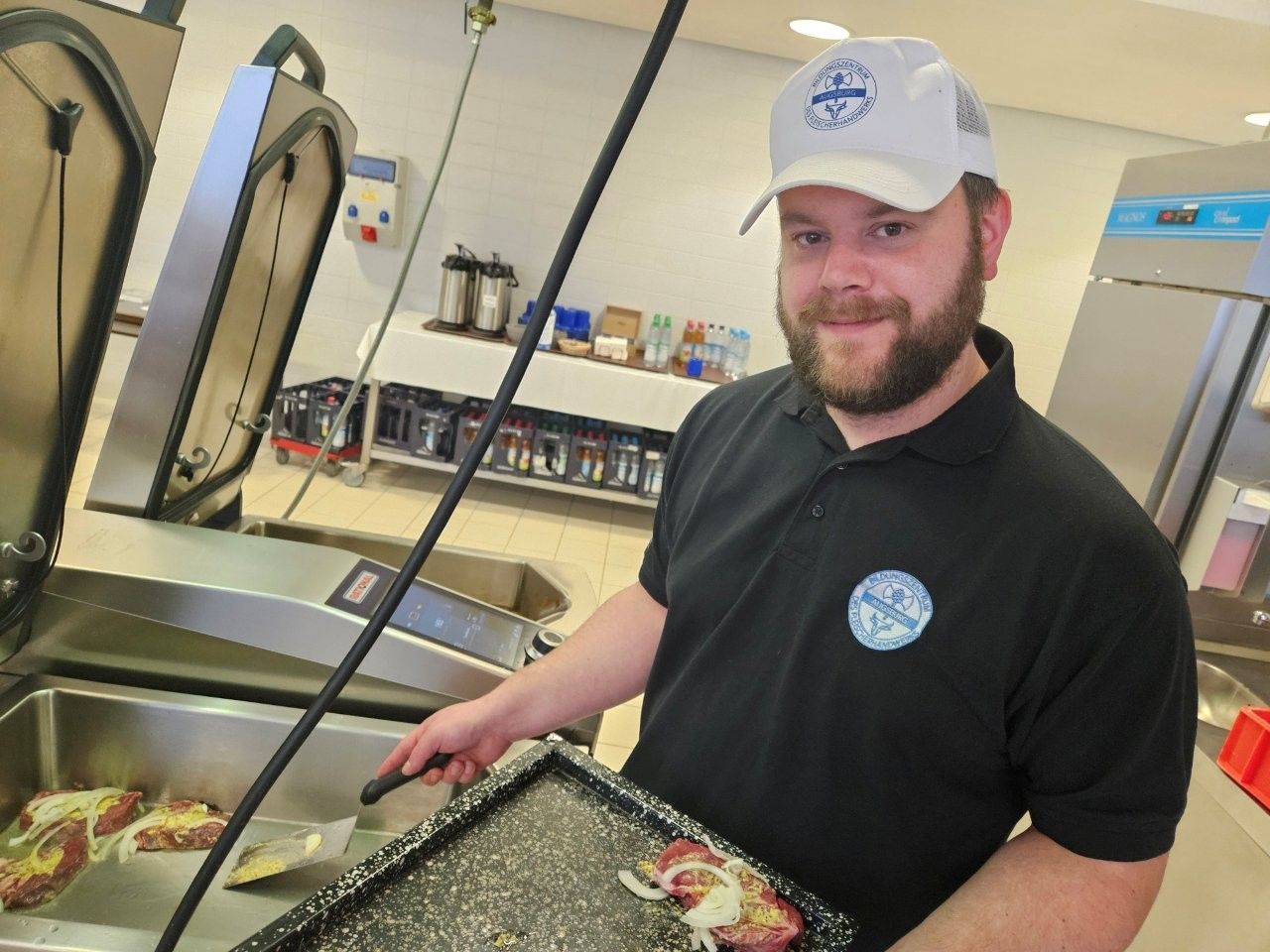 Ein Mann steht in einer Küche neben einem Küchengerät. Er hält einen Pfannenwender und ein Tablett mit Essen in der Hand. Er lächelt und trägt einen Hut.
