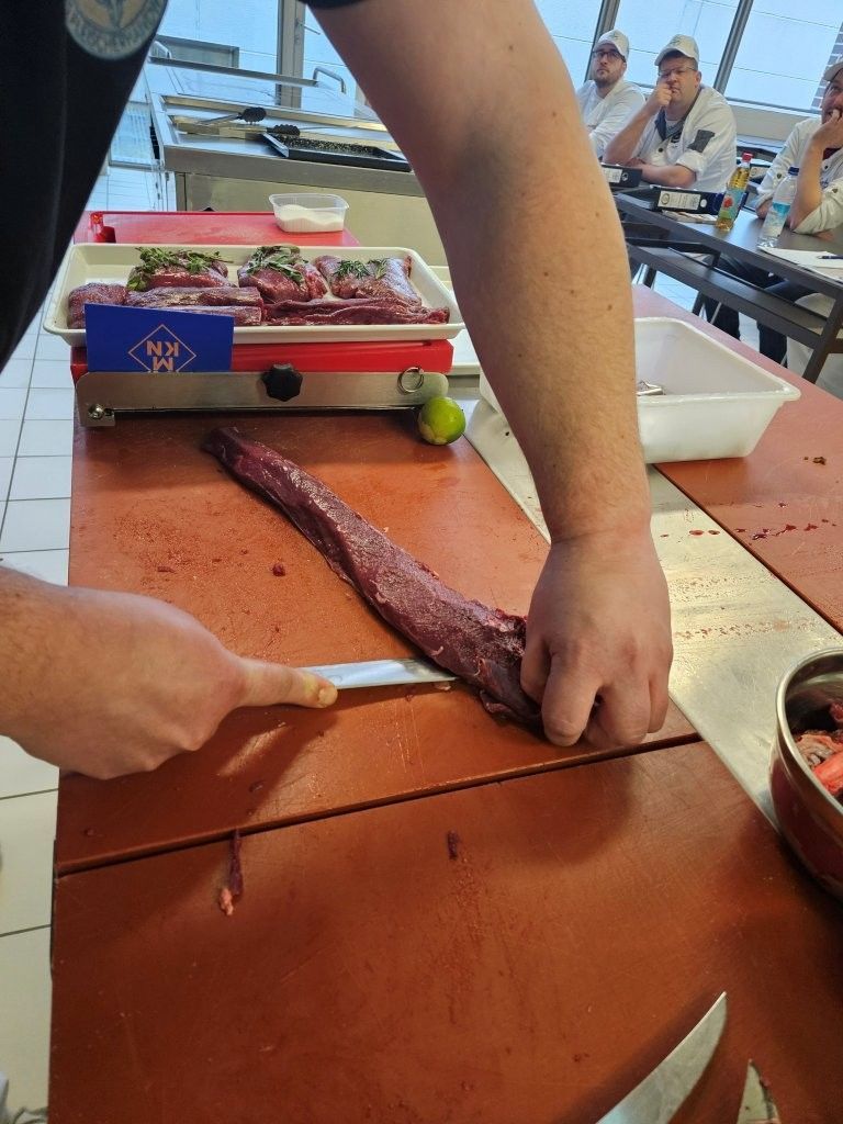 Ein Koch pariert Fleisch mit einem Messer auf einer roten Arbeitsplatte; mehrere Personen beobachten ihn im Hintergrund.