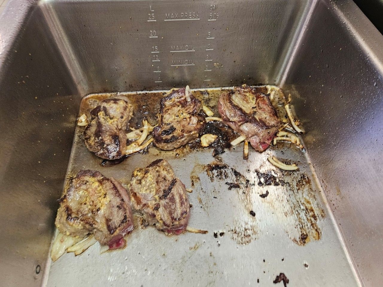 Gegrilltes Fleisch und Zwiebeln in einer Edelstahlpfanne, mit deutlichen Grillspuren und dunkelbrauner Färbung.