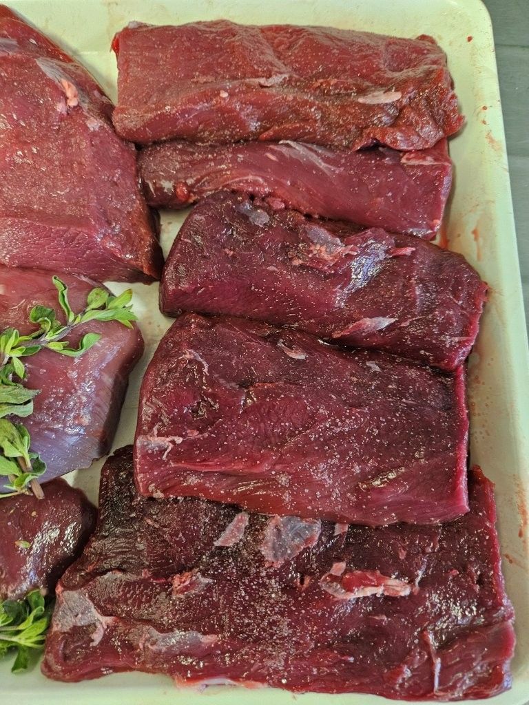 Rohe rote Fleischstücke auf einer weißen Platte, teilweise mit Salz bestreut, mit grünen Kräutern garniert.