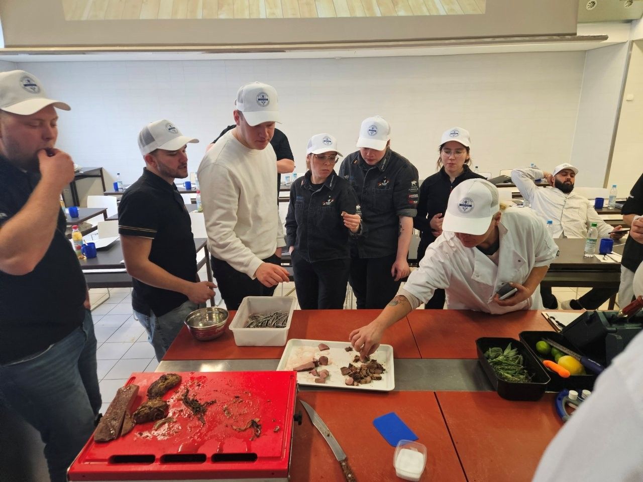 Personen mit weißen Kochmützen beobachten einen Koch bei der Zubereitung von Speisen in einer Küche.