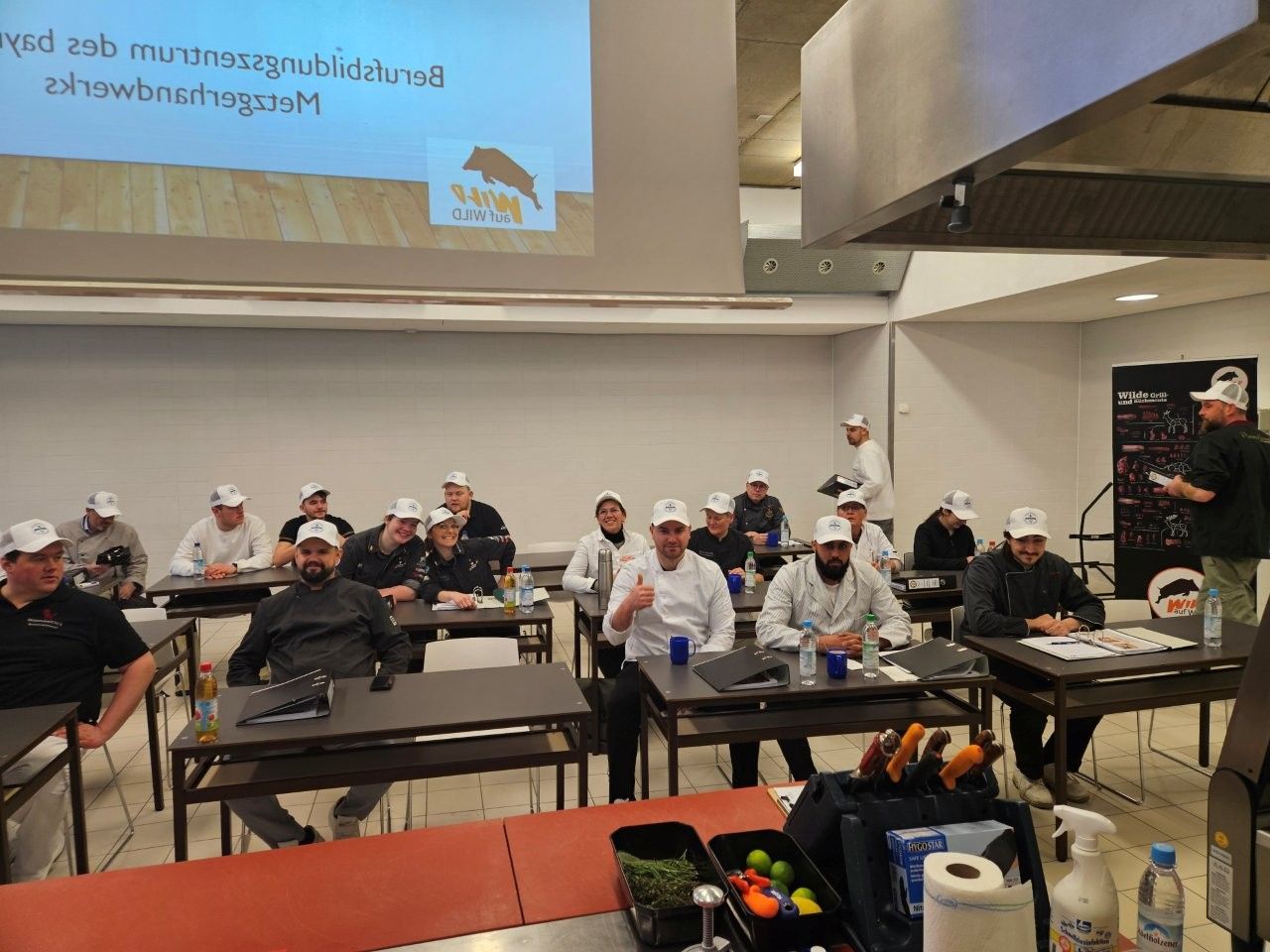 Ein Kochkurs mit Schülern, die an Tischen sitzen und eine Vorführung beobachten; alle tragen weiße Kochmützen.