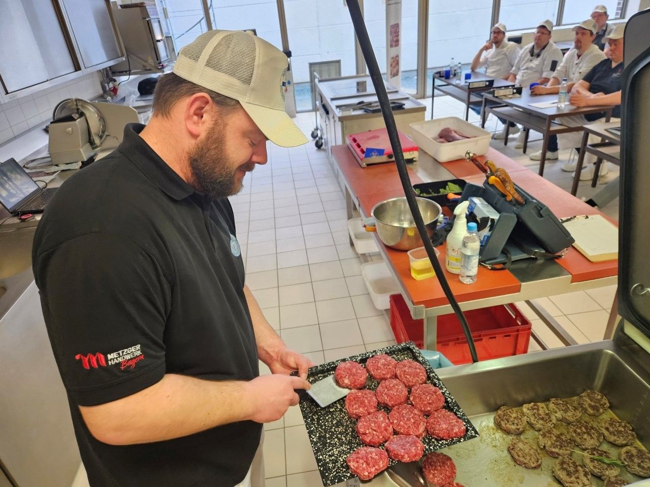 Mann im schwarzen Hemd grillt Burger, andere Personen im Hintergrund.