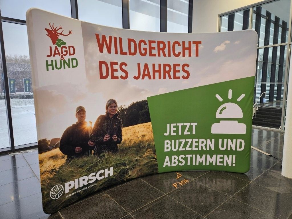 Werbeplakat für „Jagdhund Wildgericht des Jahres“. Zwei Personen auf einem Feld, grüner, weißer und roter Text, mit einem Buzzer-Symbol.