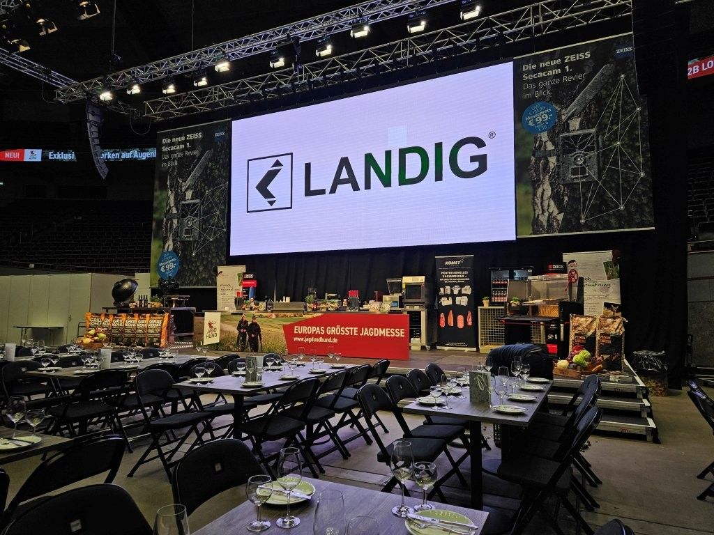 Ein Veranstaltungsraum mit Tischen, Stühlen und einem Bildschirm, auf dem das Logo „LANDIG“ angezeigt wird.