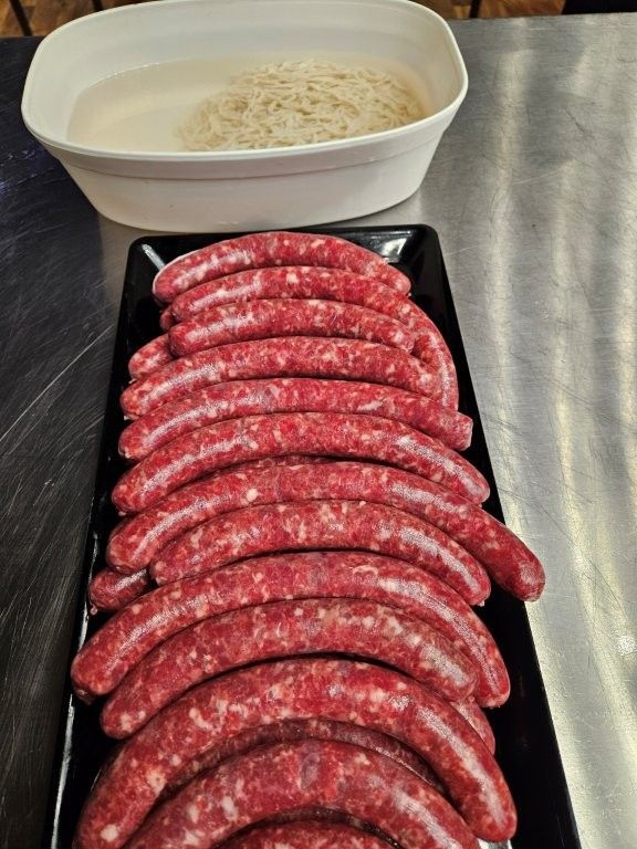 Rohe Würstchen auf einem schwarzen Tablett, im Hintergrund eine Schüssel mit Nudeln.