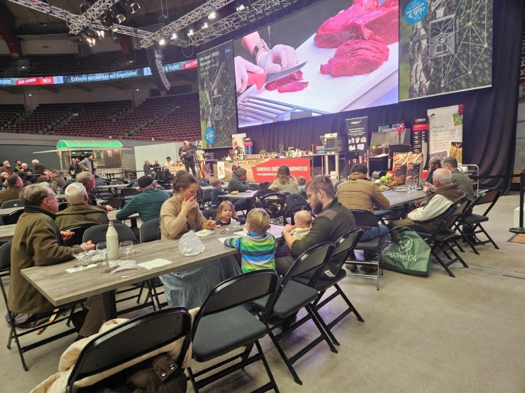 Menschen sitzen an Tischen und verfolgen eine Kochvorführung auf einer Großleinwand bei einer Veranstaltung in einem großen Veranstaltungsort.