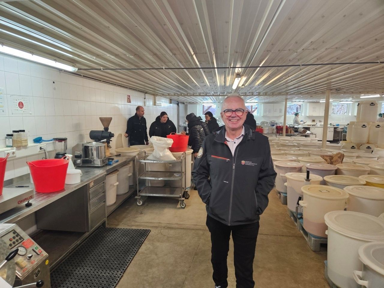 Ein Mann in Jacke lächelt in einer Lebensmittelproduktionsanlage, umgeben von Personal und Ausrüstung.