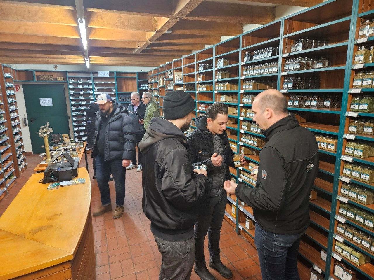 Menschen stöbern in Regalen mit Einmachgläsern in einem Laden, möglicherweise einer Apotheke. Ein Mann unterhält sich mit zwei anderen.