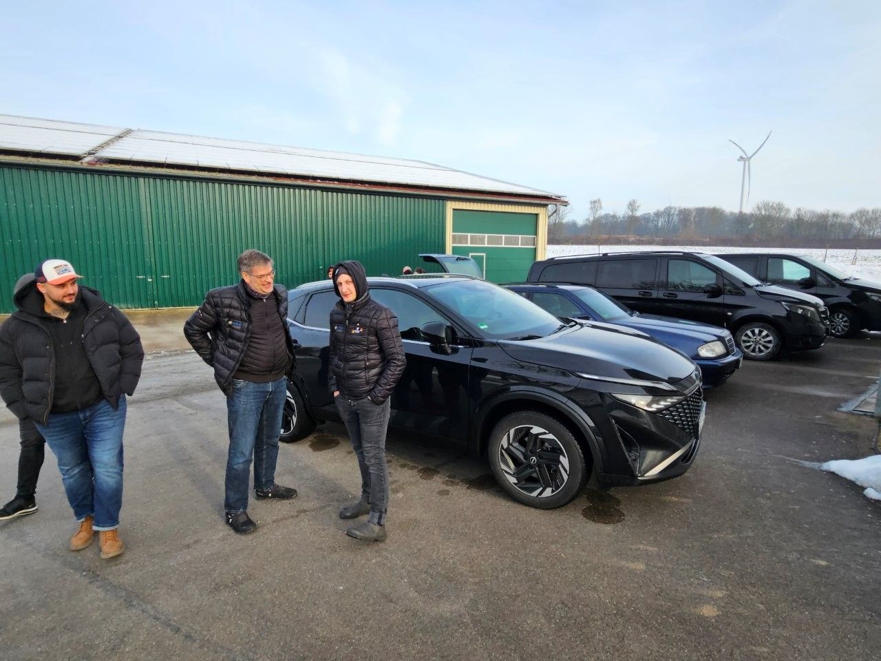 Drei Männer stehen neben einem schwarzen Geländewagen und anderen Fahrzeugen an einem grünen Gebäude und einem schneebedeckten Feld.
