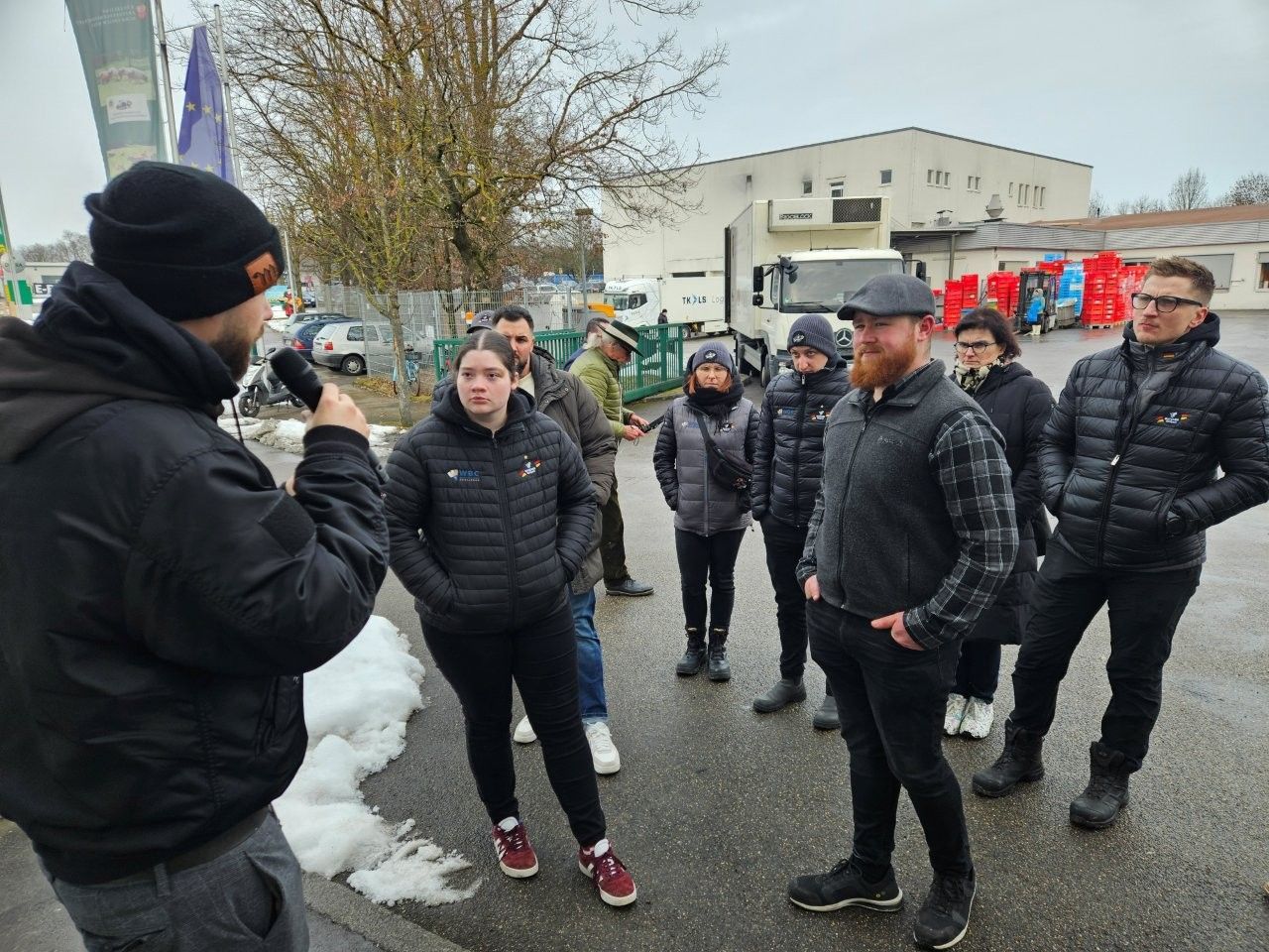 Eine Gruppe von Menschen hört einem Mann zu, der im Freien spricht. Einige tragen Mäntel und stehen in der Nähe von Gebäuden und Fahrzeugen.