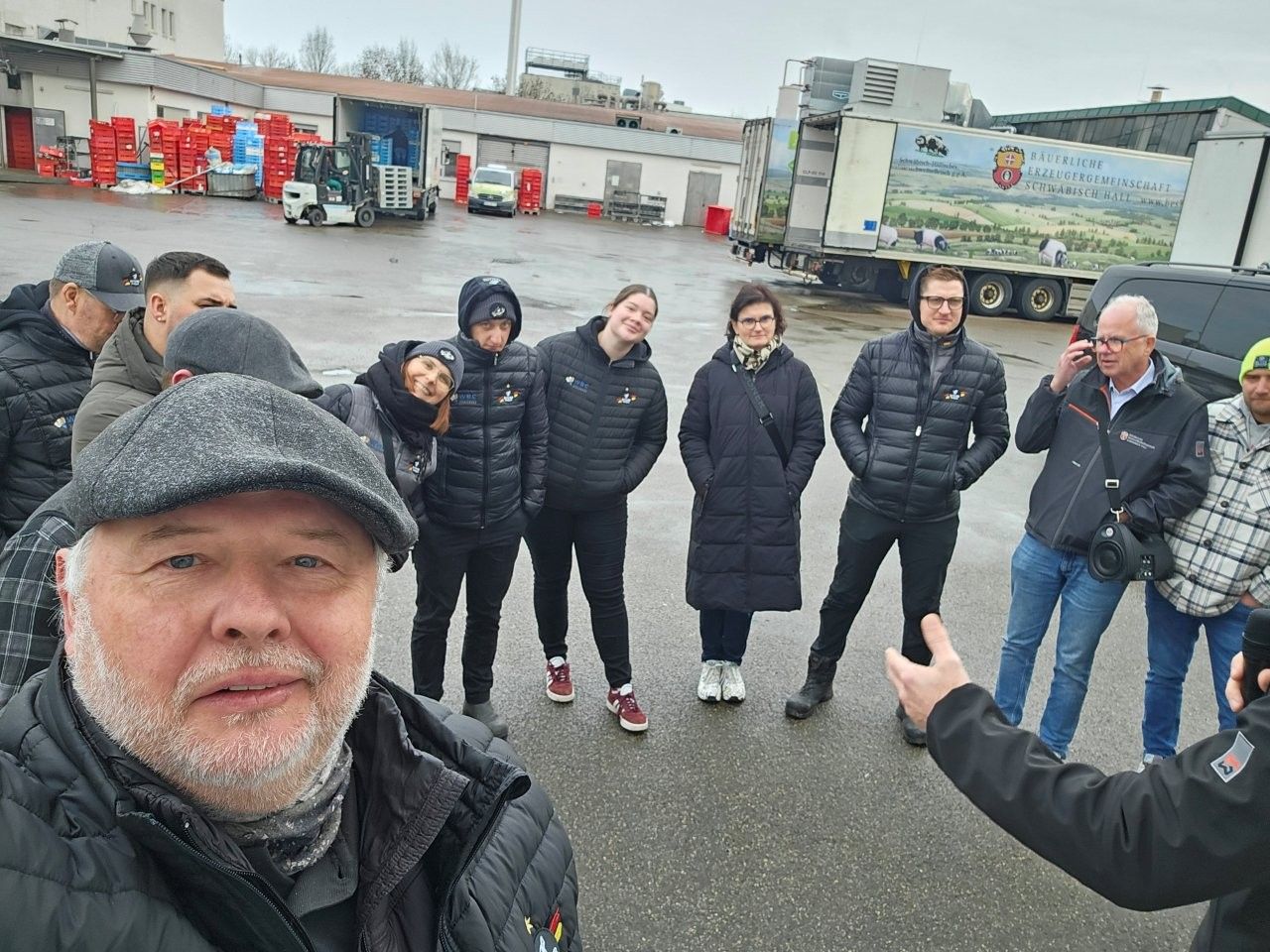 Eine Gruppe von Menschen steht im Freien vor einer Fabrik, einige schauen in die Kamera. Bewölkter Himmel.