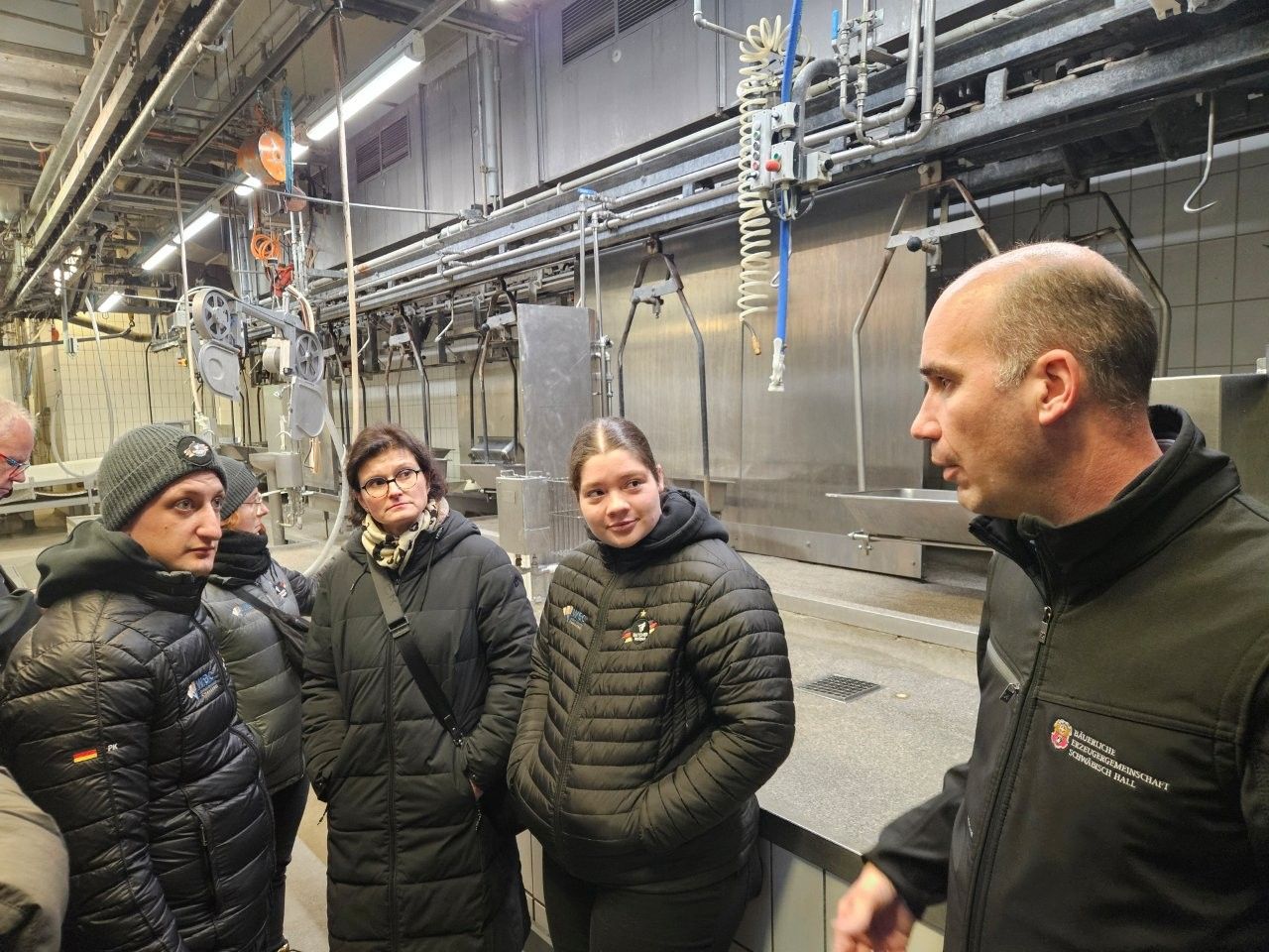 Eine Gruppe von Menschen besichtigt eine moderne Industrieanlage und hört einem Mann in einer Weste zu.