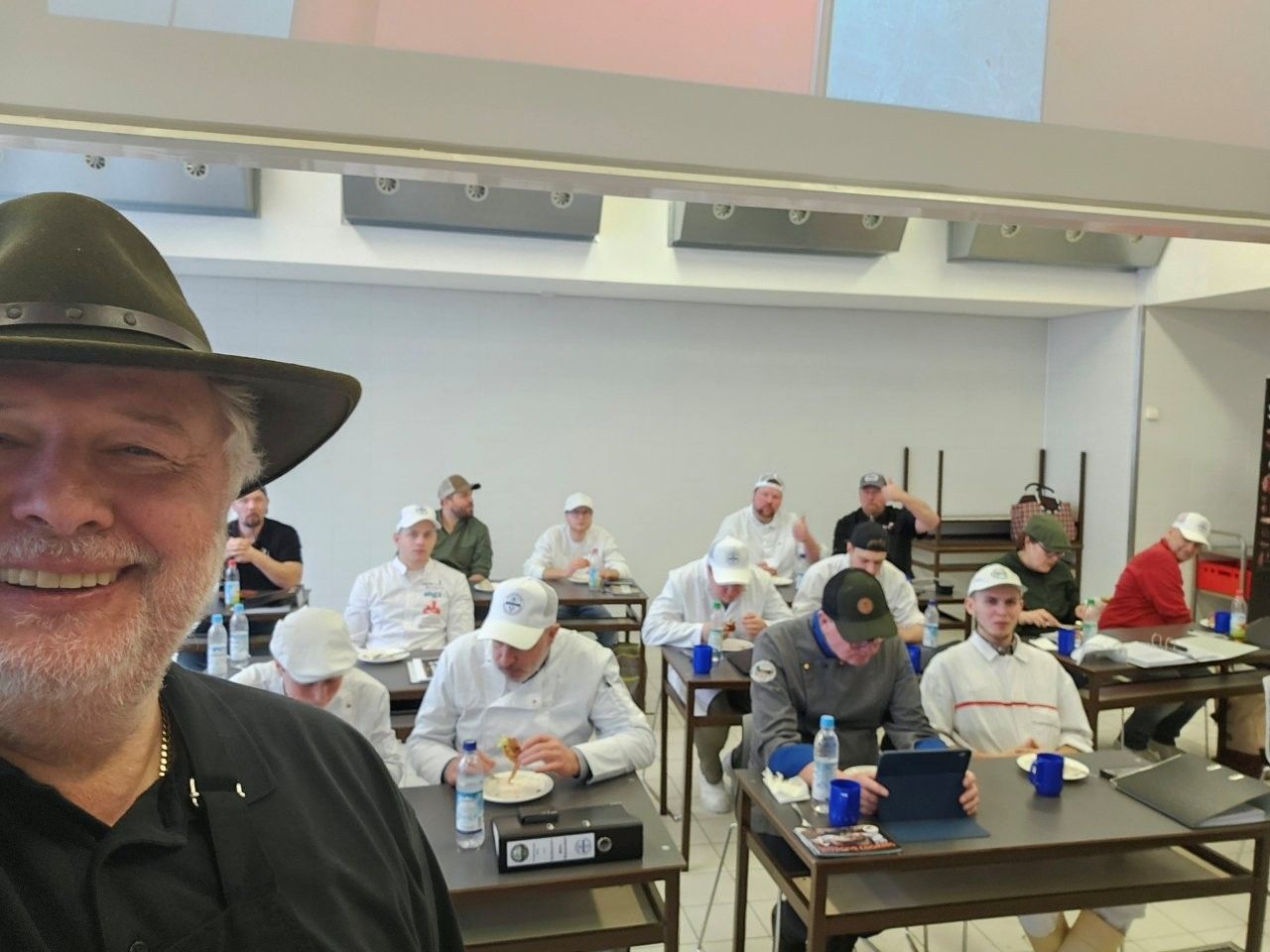 Mann mit Hut macht ein Selfie in einem Klassenzimmer mit Schülern an ihren Tischen.