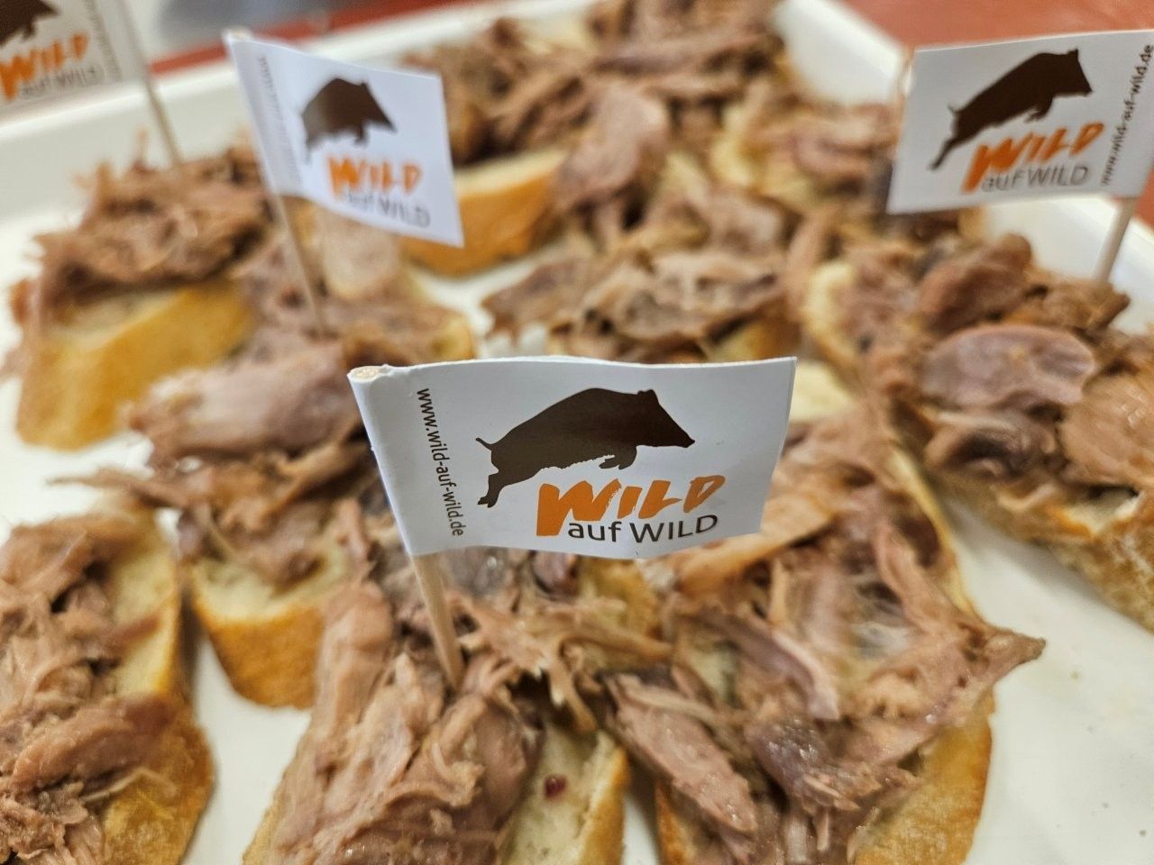 Tapas mit zerkleinertem Fleisch, etikettiert als „Wild but Wild“, serviert auf Baguettescheiben auf einem Tablett.