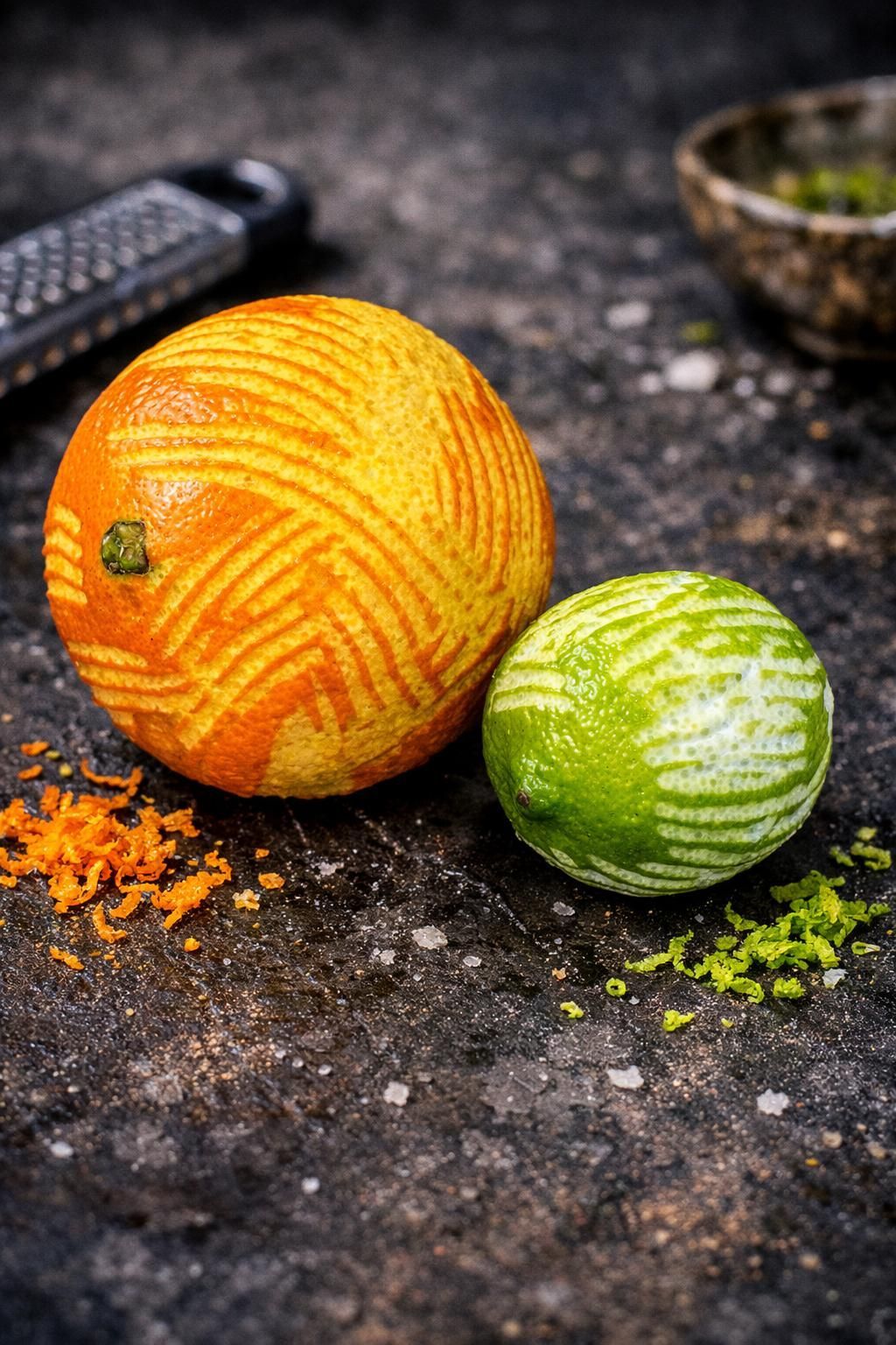 Orangen- und Limettenzesten mit Abriebmustern neben einer Reibe und einer Schale mit Zitronenschale auf einer dunklen Oberfläche.