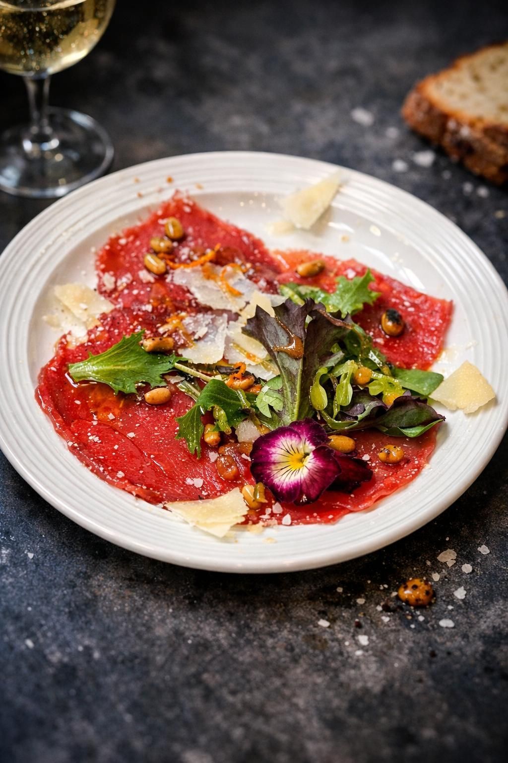 Rindercarpaccio mit Rucola, Pinienkernen, Parmesan und einer Blüte auf einem weißen Teller.
