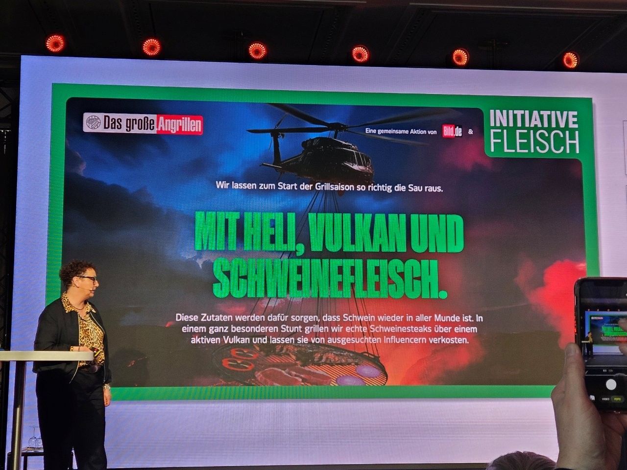 Eine Frau präsentiert ein Plakat mit der Aufschrift „Mit Hölle, Vulkan und Schweinefleisch“. Hubschraubergrafik, grüne und rote Farben.