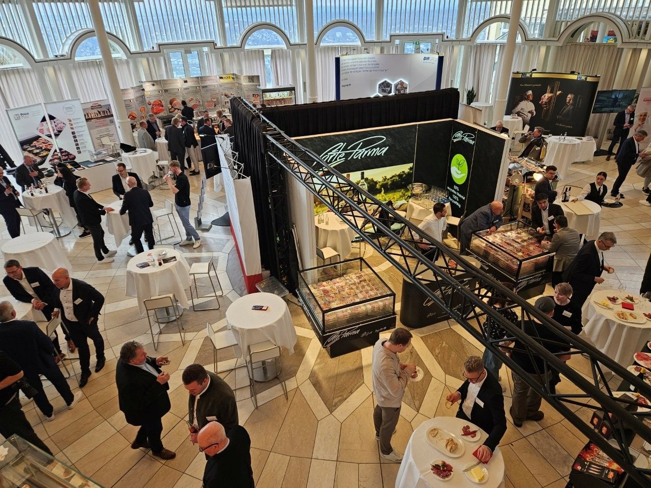 Großer Veranstaltungssaal mit Teilnehmern; Stände, Tische und eine Traversenkonstruktion sind aufgebaut. Der Raum ist lichtdurchflutet.