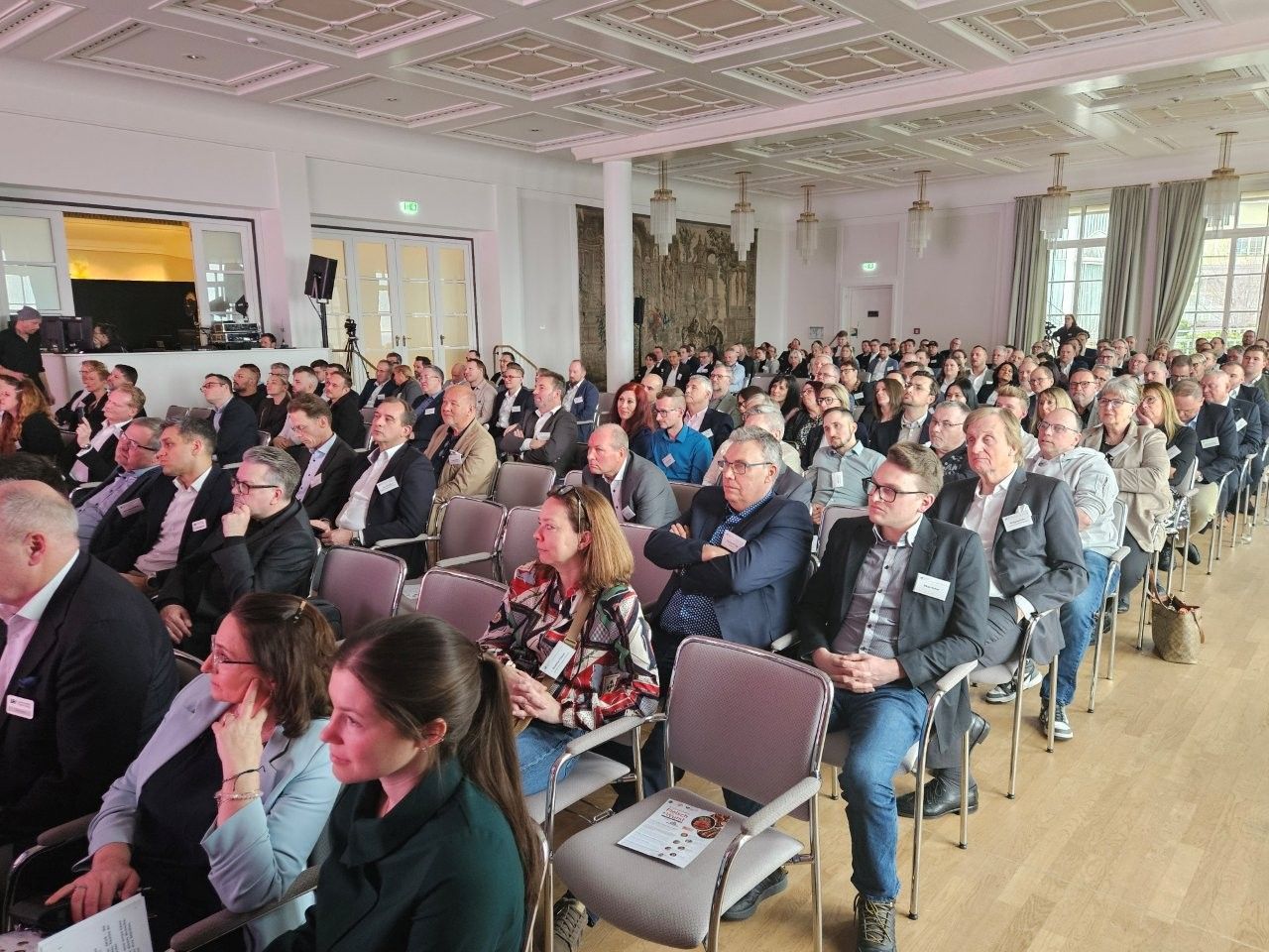 Großer Konferenzsaal voller Teilnehmer, die einem Redner zuhören. Neutrale Beleuchtung.