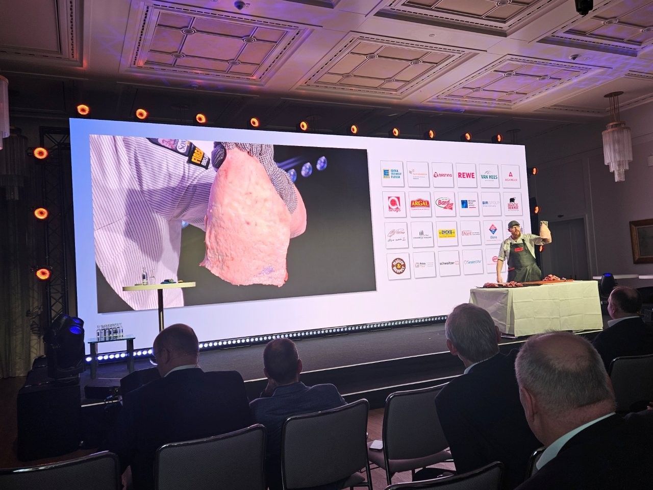 Ein Moderator demonstriert auf der Bühne das Zerlegen von Fleisch; die Demonstration wird auf eine große Leinwand projiziert, während ein Publikum zuschaut.